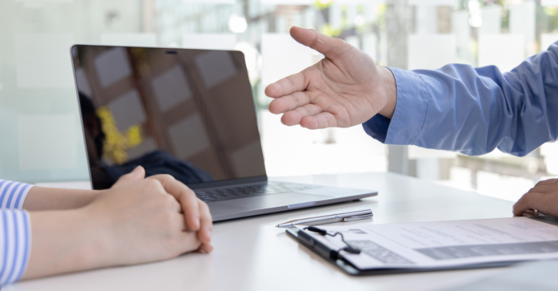 Photographie d'un entretien d'embauche, entre deux personnes, avec un ordinateur portable ouvert sur la table, un CV sur une tablette à pince et une main tendue illustrant une discussion active.